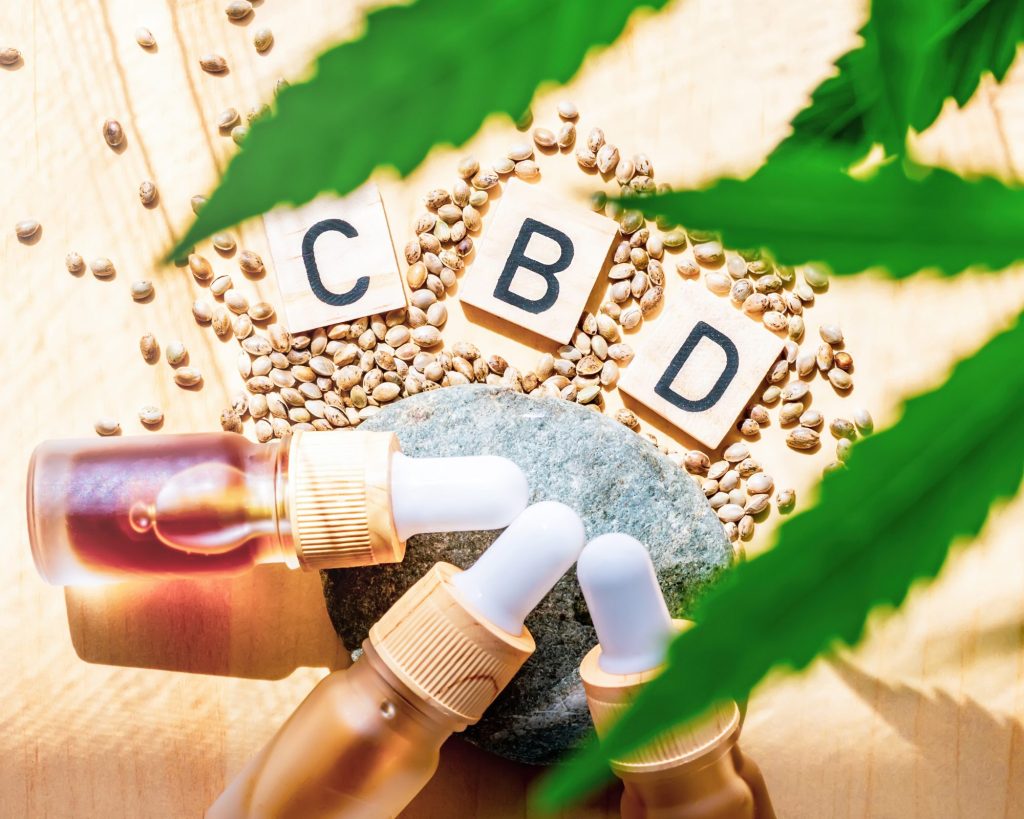olejki cbd leżące obok ziaren konopnych i liści. Nad nimi jest napis cbd ułożony z kliocków scrable